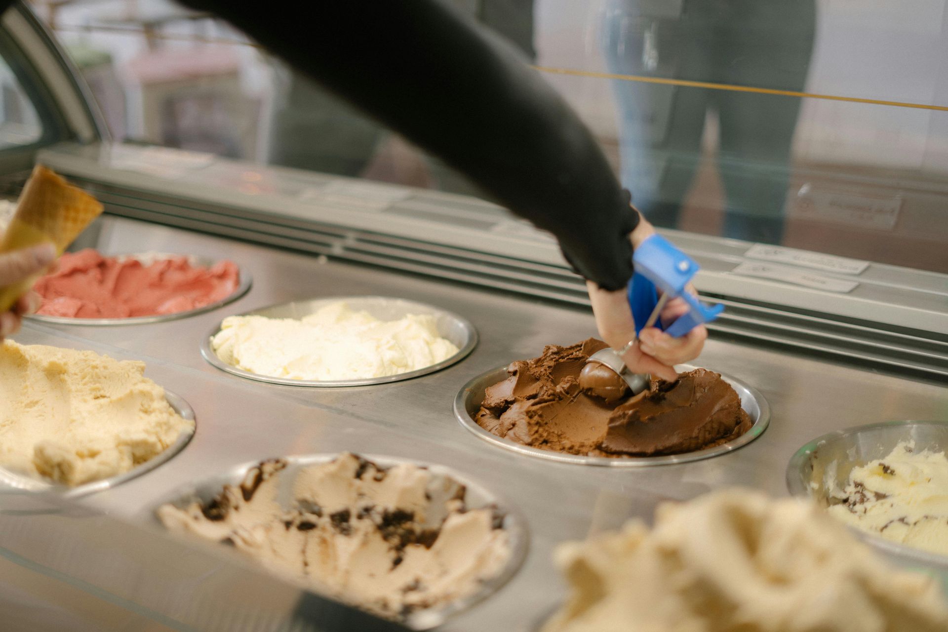 Hand using a blue scoop to serve chocolate ice cream from a shop display case
