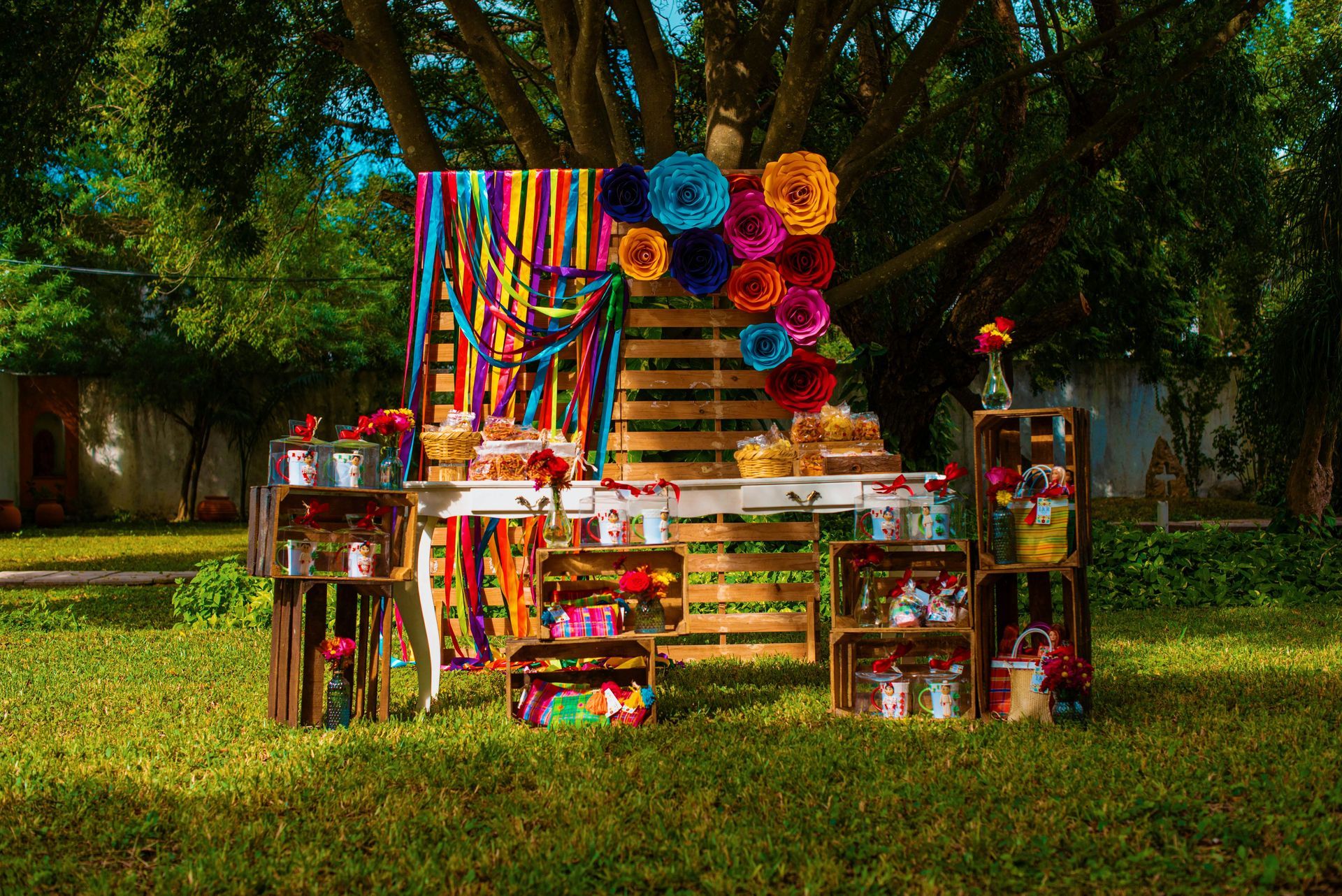 Colorful outdoor market stall with rainbow textiles, crafts, and flowers under large trees