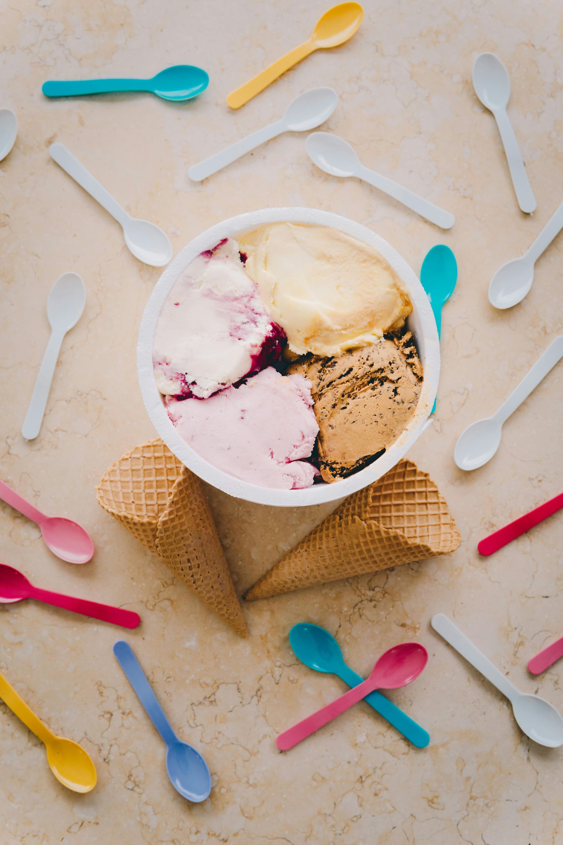 Ice cream in a cone topped with scoops, surrounded by colorful plastic spoons on a beige surface
