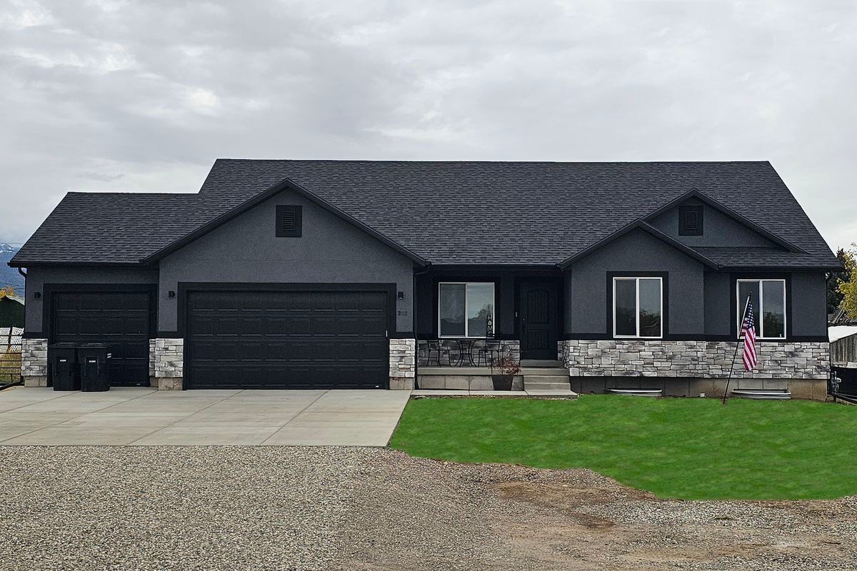 Dark gray single-story house with stone accents, three garage doors, and a gravel driveway under cloudy skies