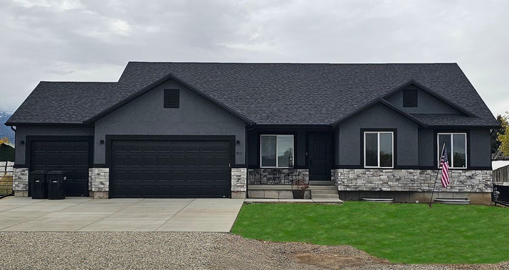 Modern black suburban house with stone accents, three-car garage, and a green lawn under a cloudy sky