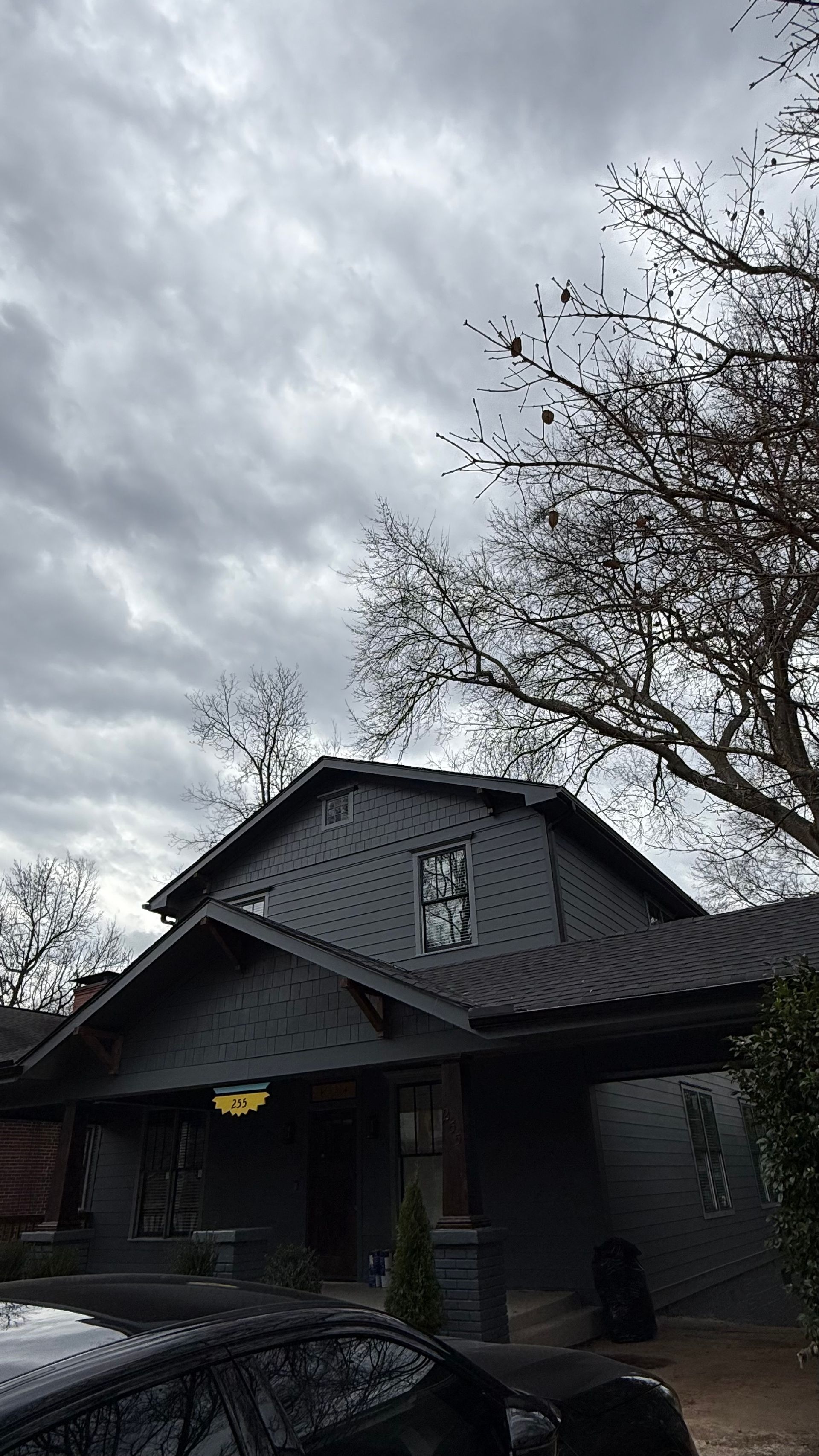 Dark house with a steep roof under a cloudy sky, framed by bare trees and a parked car in front.