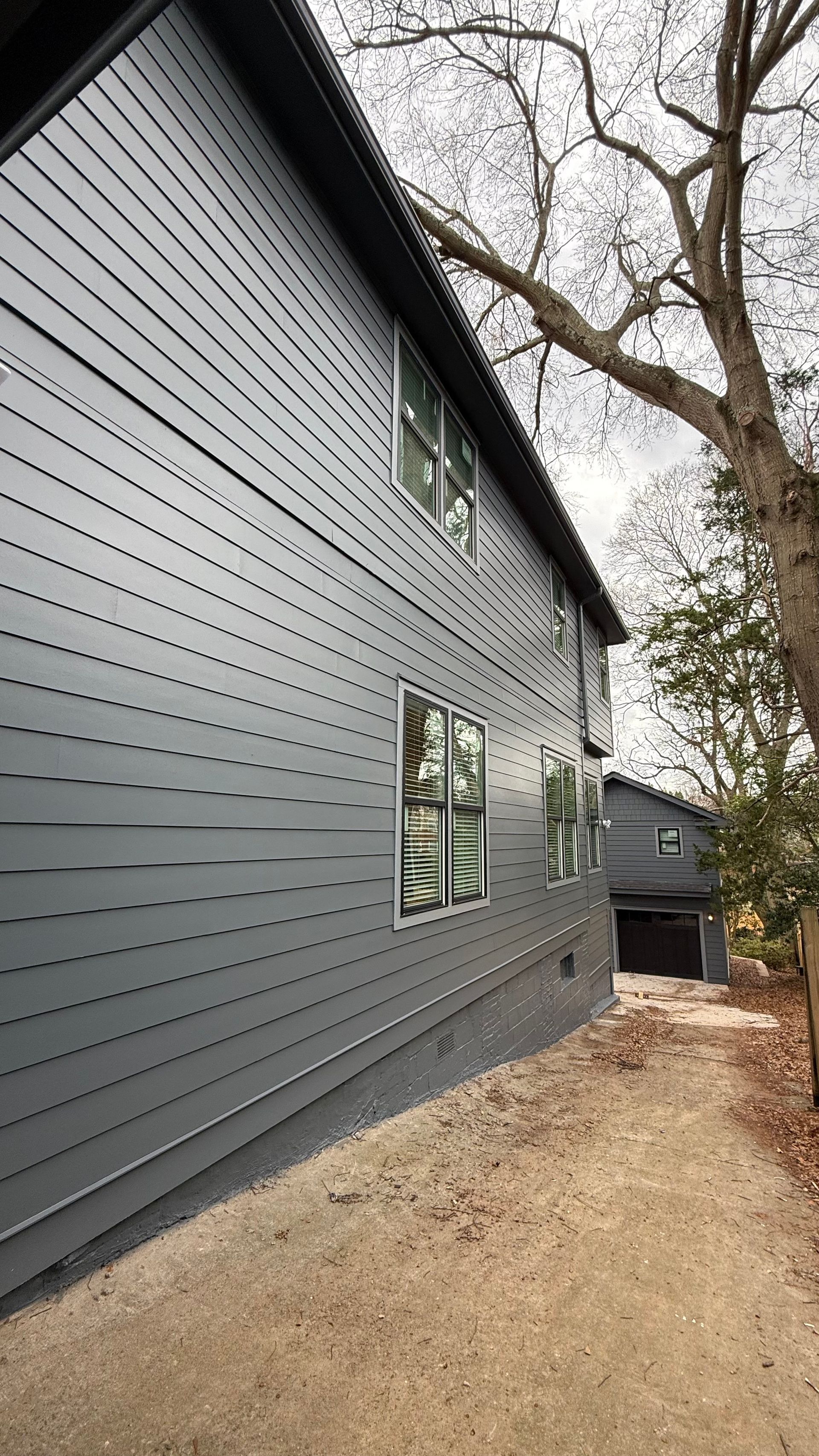 Gray house siding with windows and a dirt path beside bare trees and another house in the background