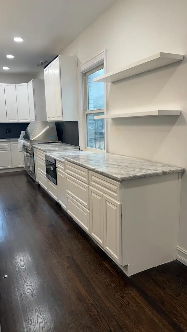 Bright narrow kitchen with white cabinets, marble counters, dark wood floors, and floating shelves by a window.