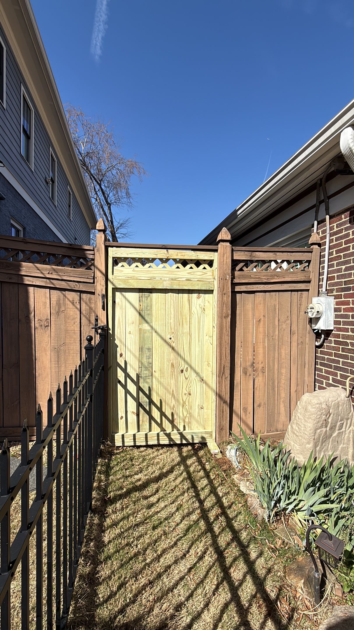 Narrow brick alley with a wooden gate, fence shadows, and blue sky overhead