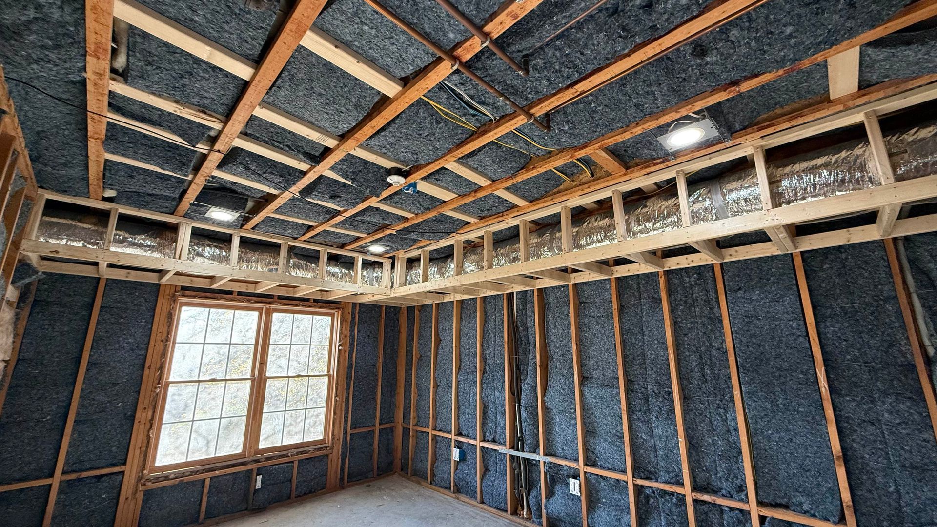 Interior of a room under construction with exposed wood framing, insulation, and a window on the left.