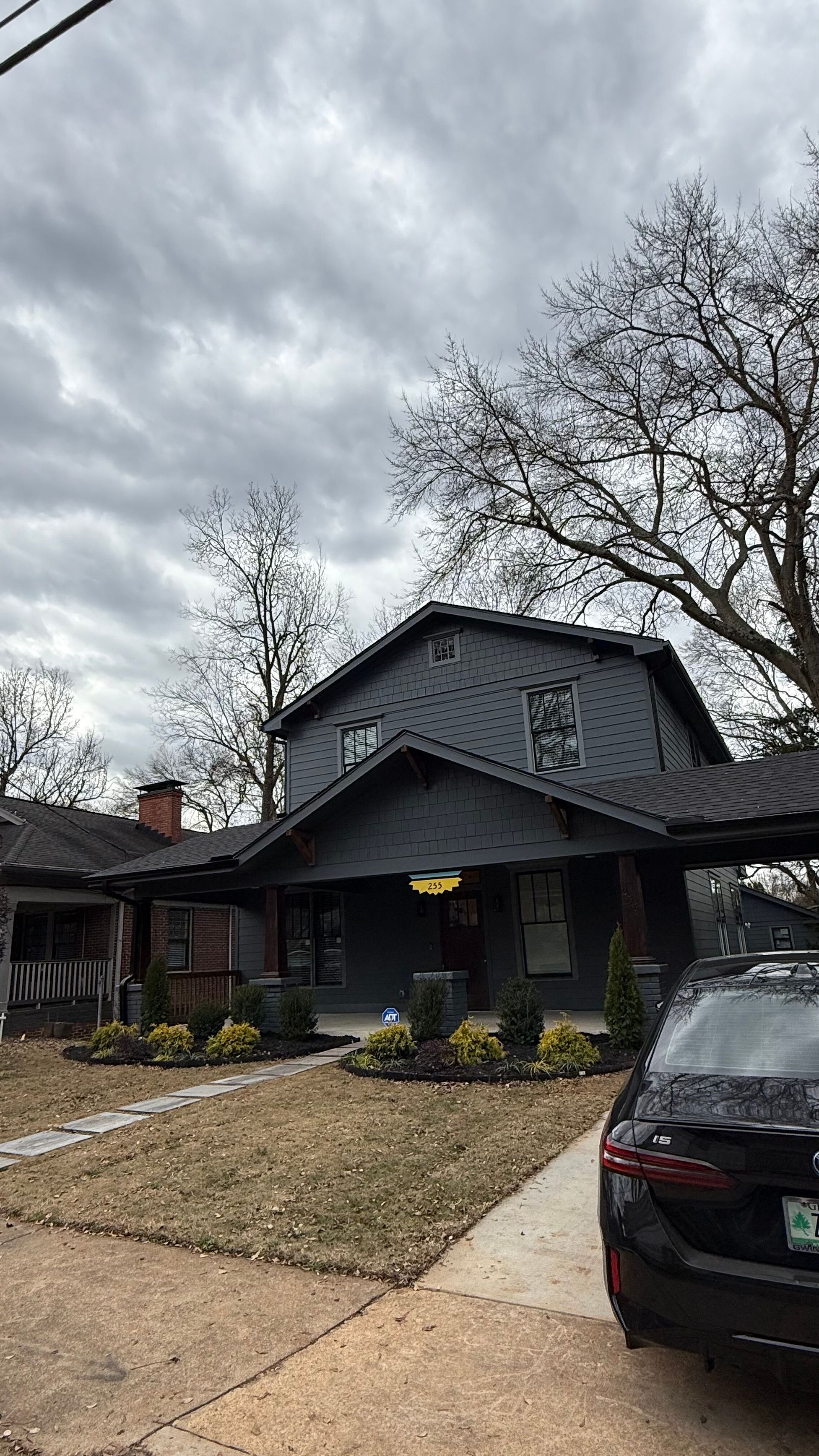 Two-story house with dark roof and porch, front yard path, and a black car parked in the driveway under cloudy sky