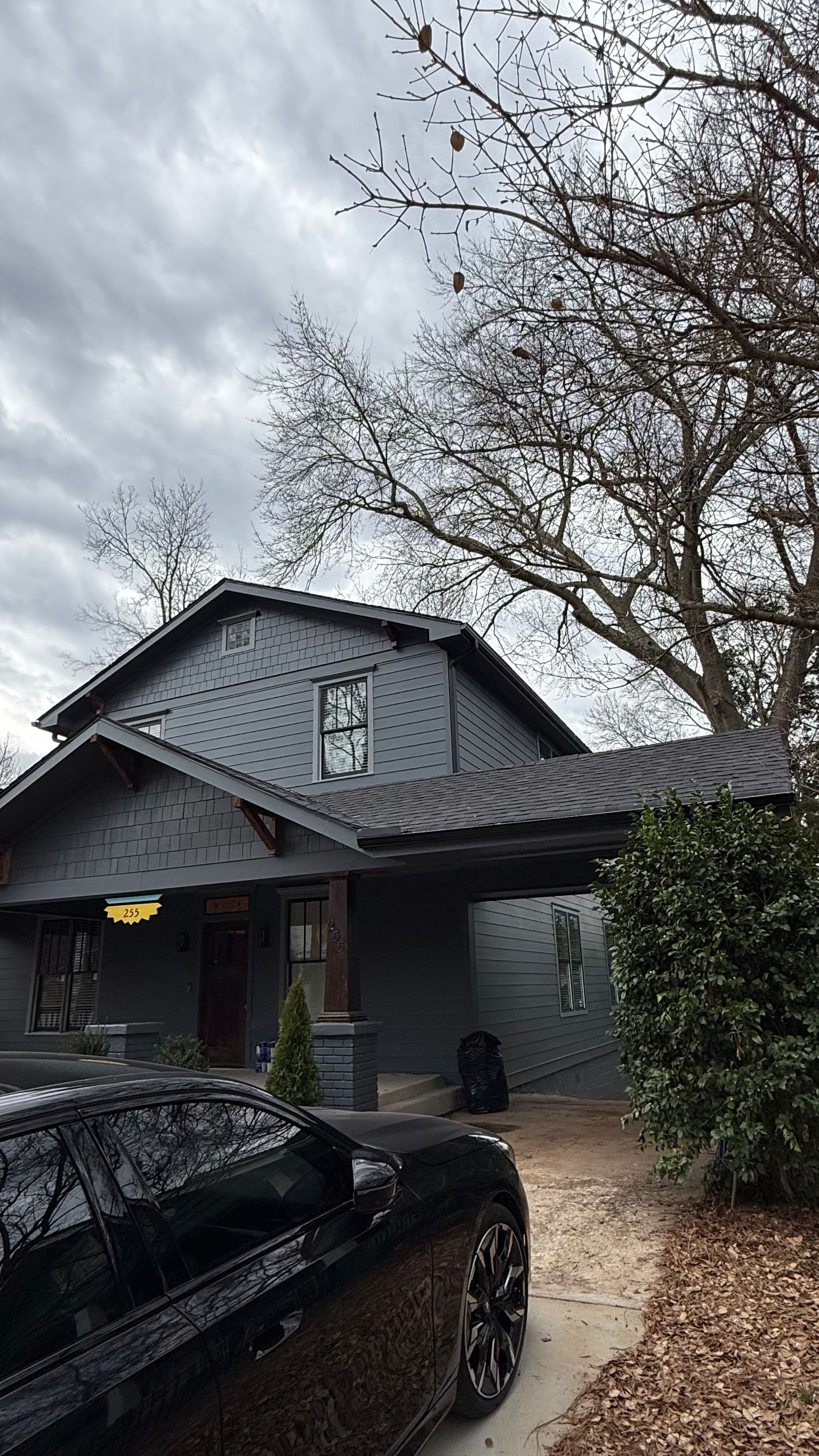 Dark gray house with a black car in front under cloudy skies, lined by trees and shrubs.