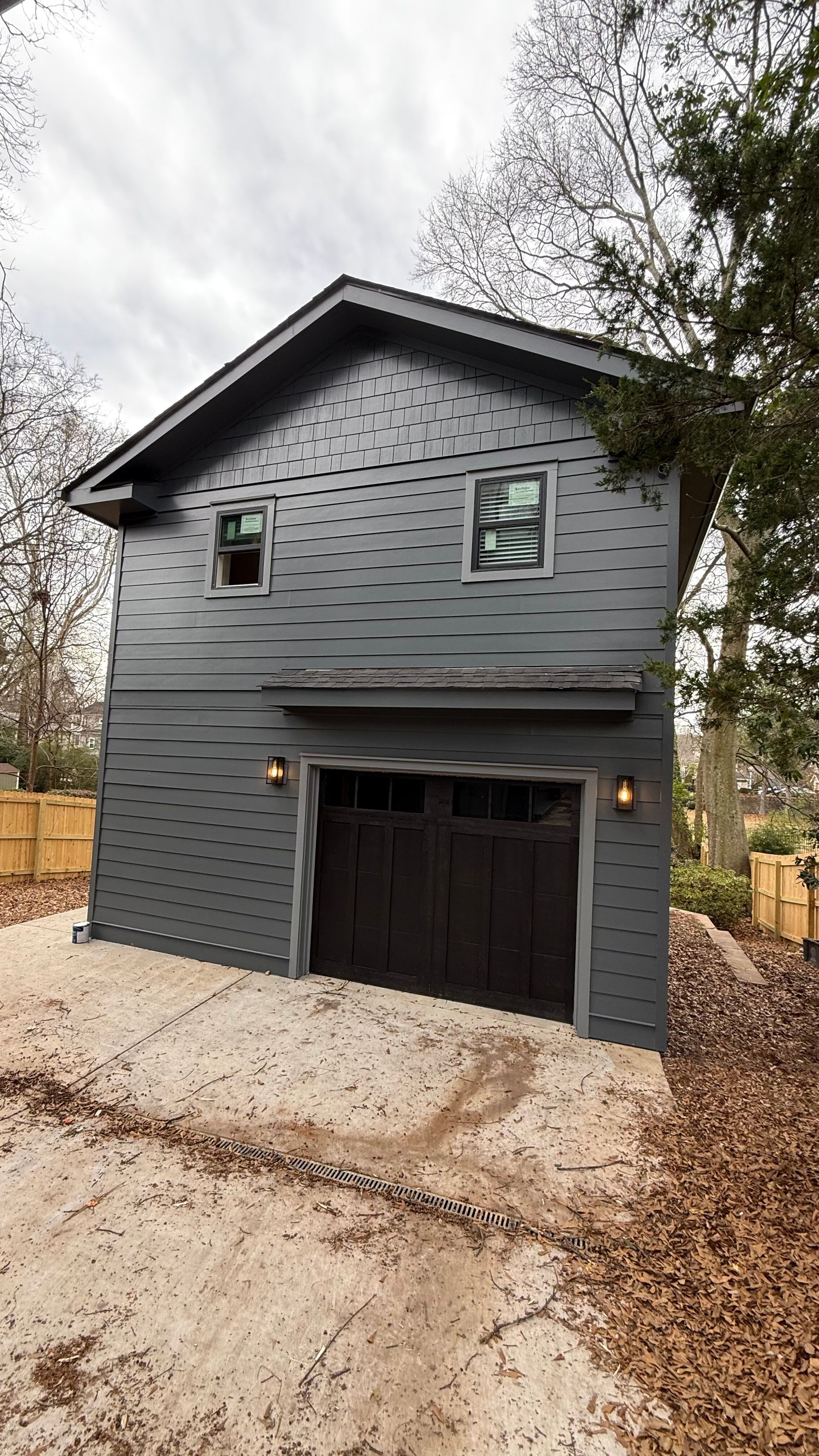Gray two-story garage or small house with dark trim, a black door, and leaf-covered driveway in a wooded area
