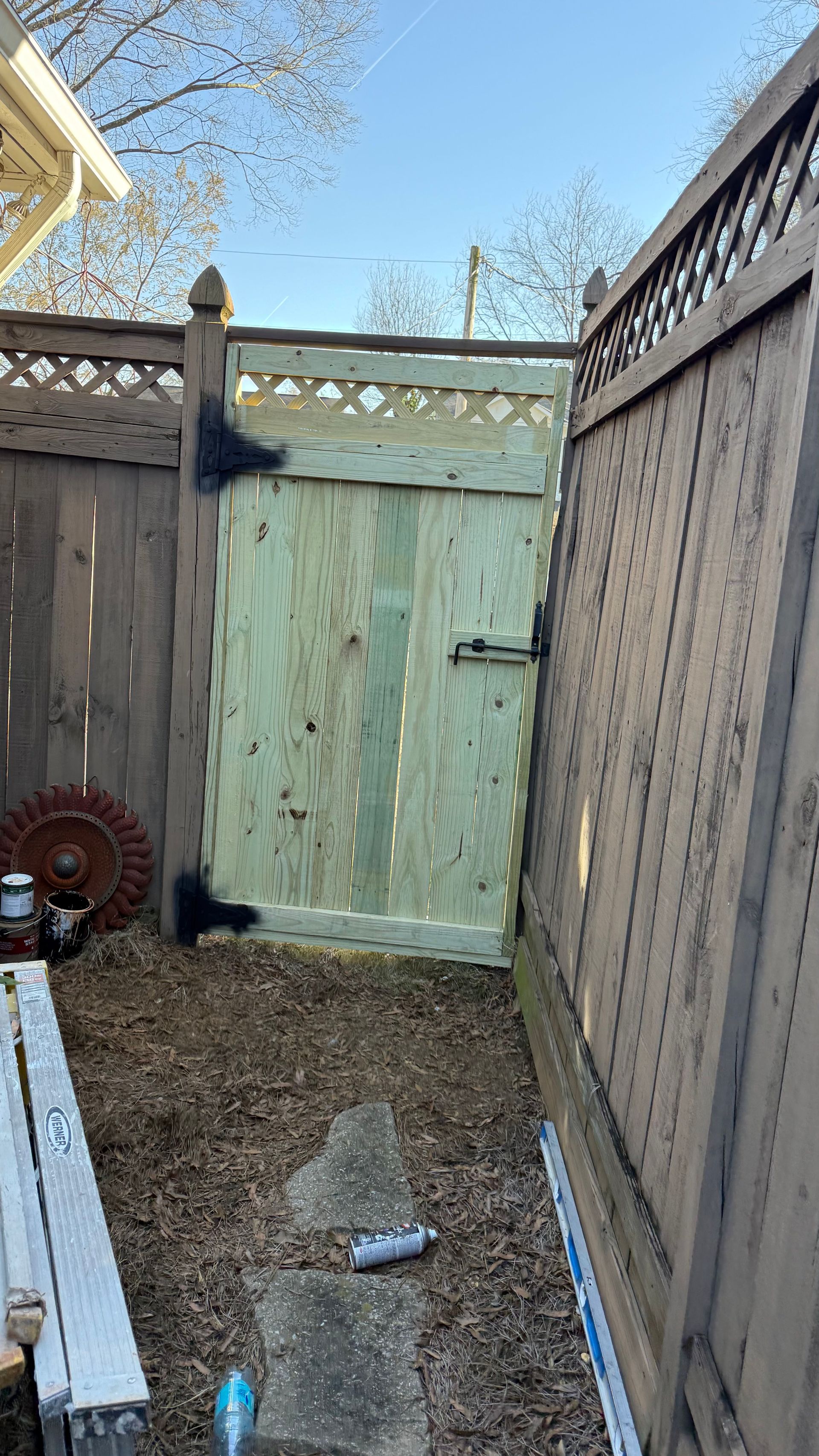Narrow gravel alley with weathered wooden fences and a closed green gate under a blue sky