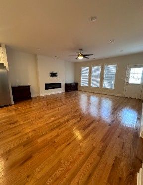 Empty living room with glossy hardwood floors, ceiling fan, fireplace, and shuttered windows.