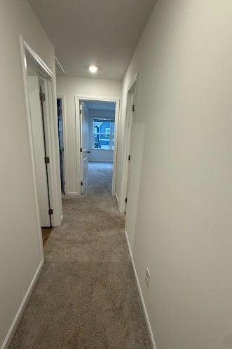 Narrow white hallway with beige carpet and doors, leading to a bright room at the end