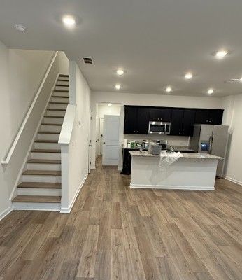 Modern open-concept kitchen and living area with wood floors, white walls, dark cabinets, and a staircase.