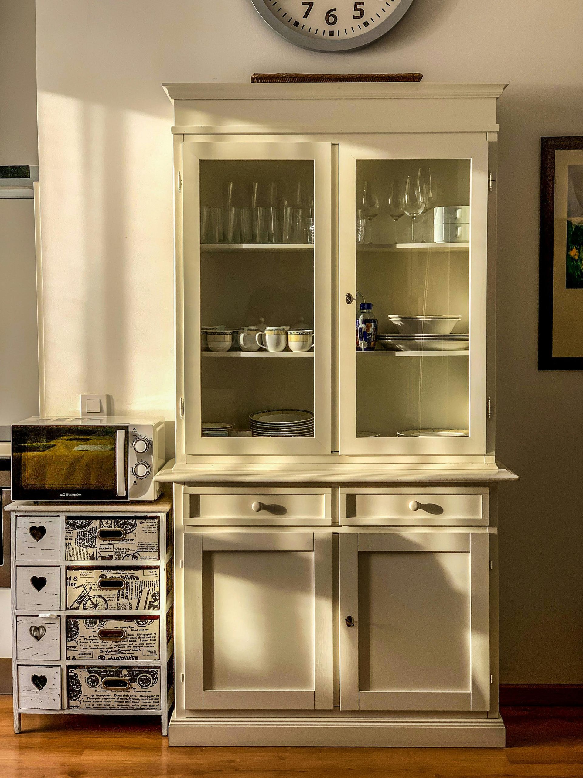 White china cabinet with glass doors and drawers beside a small patterned storage chest in a sunlit room.