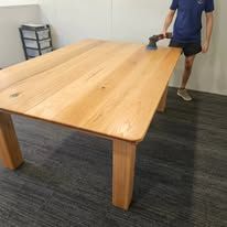 Person sanding a large wooden table in a room with dark carpet