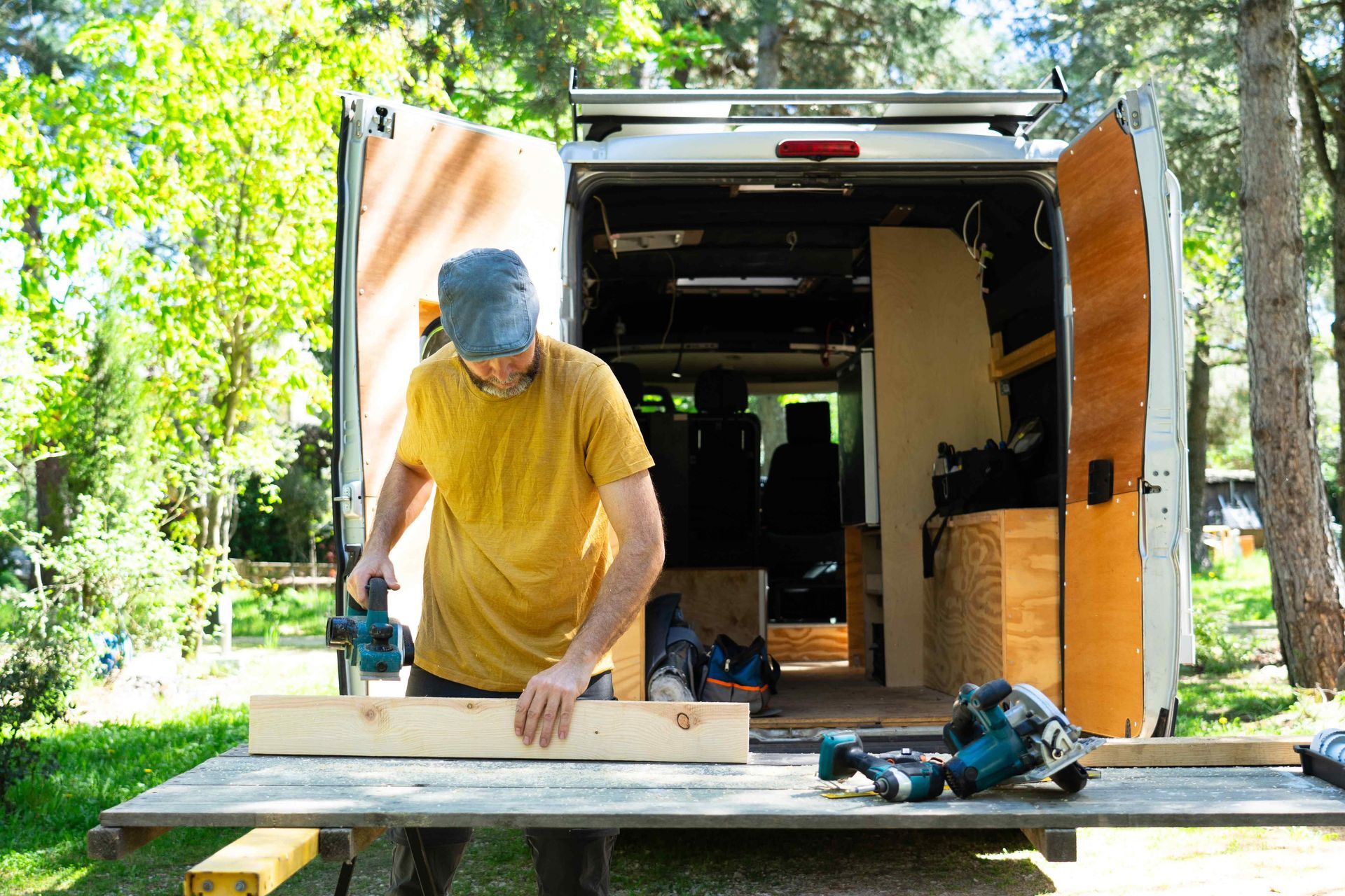 Person in a yellow shirt working on a wooden board at a table outside a van in a wooded area