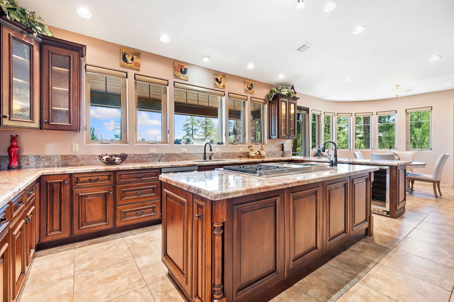 Spacious kitchen with dark wood cabinets, marble island, large windows, and beige tile floor.