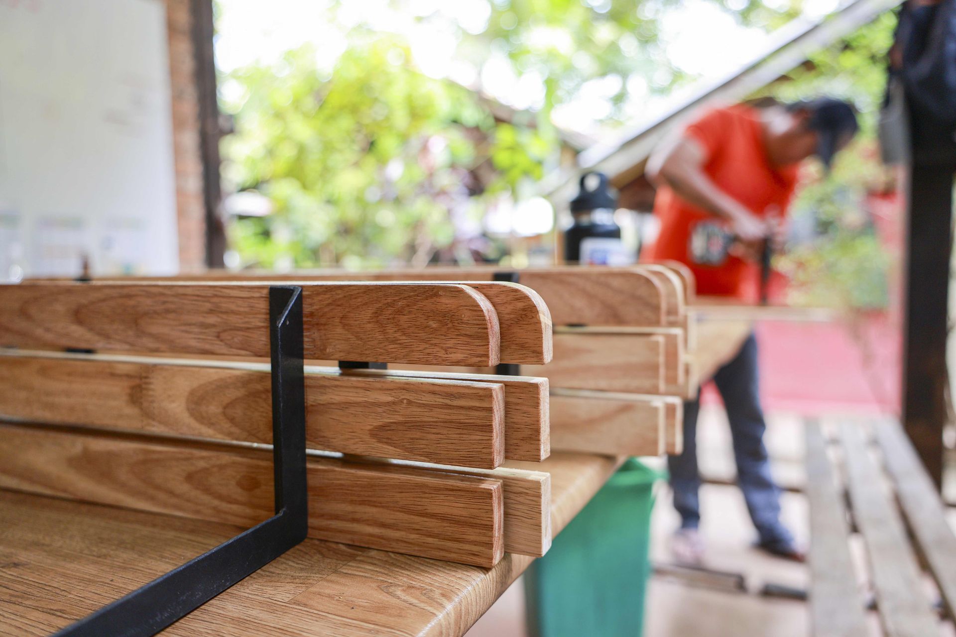 Stacked wooden boards with black metal brackets, with a person working blurred in the background.