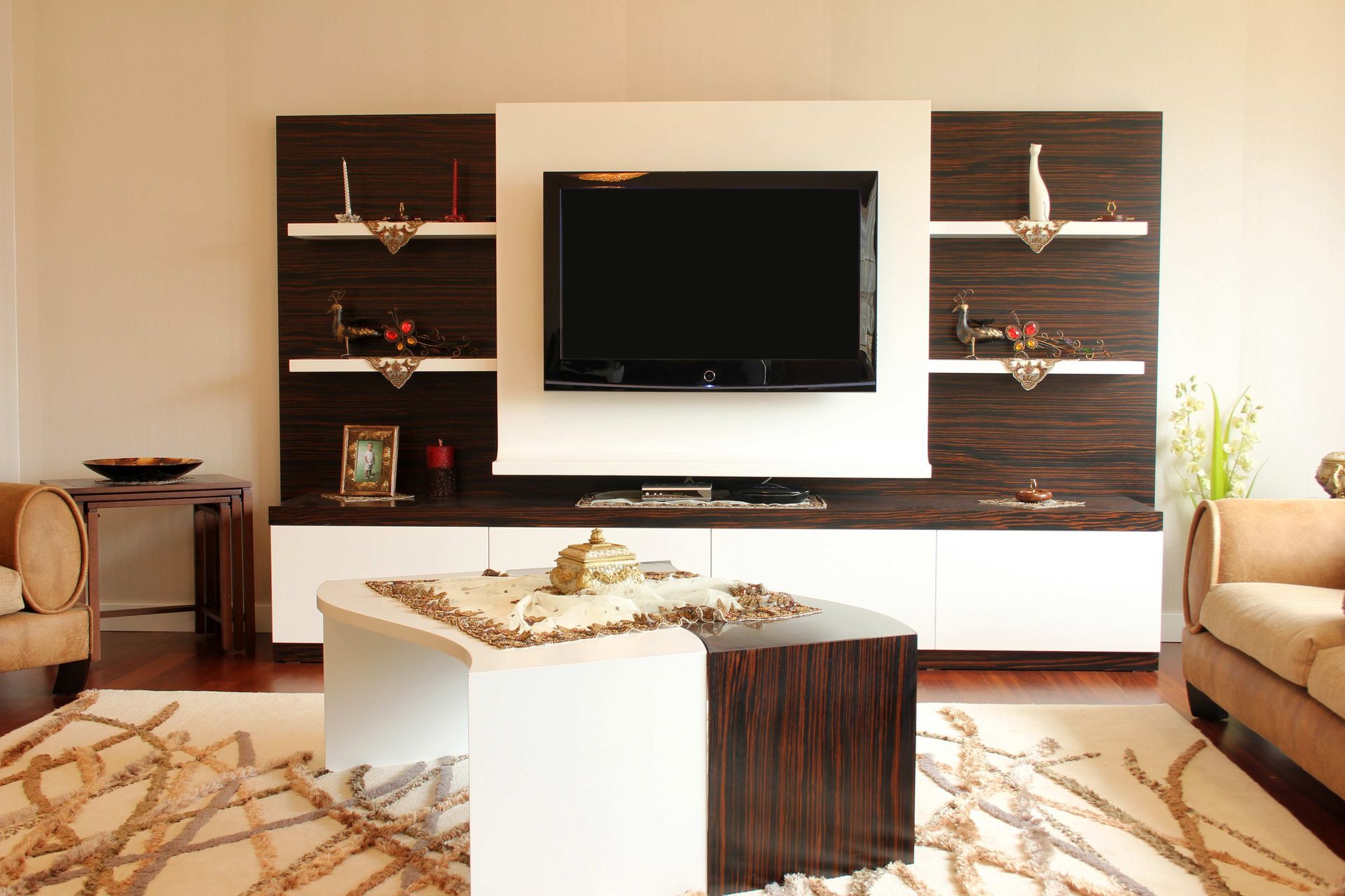 Modern living room with wall-mounted TV, dark wood shelves, white media console, and beige sofas