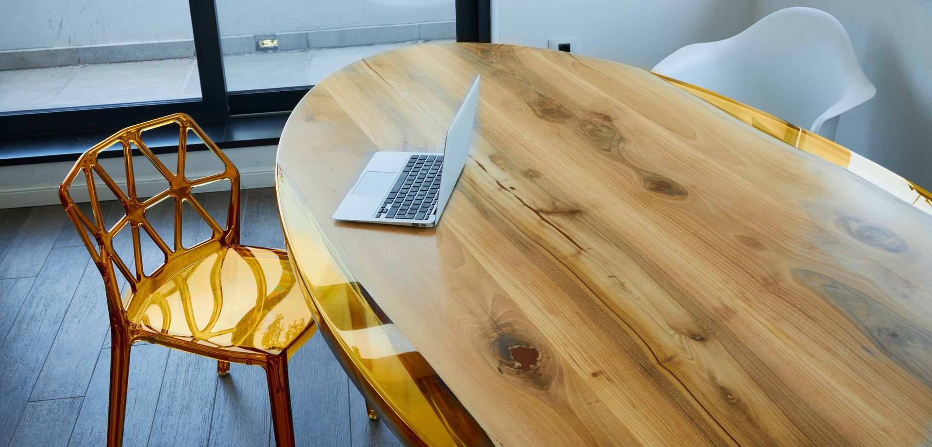 Wooden oval table with a laptop, flanked by a yellow chair and a white chair in a bright room