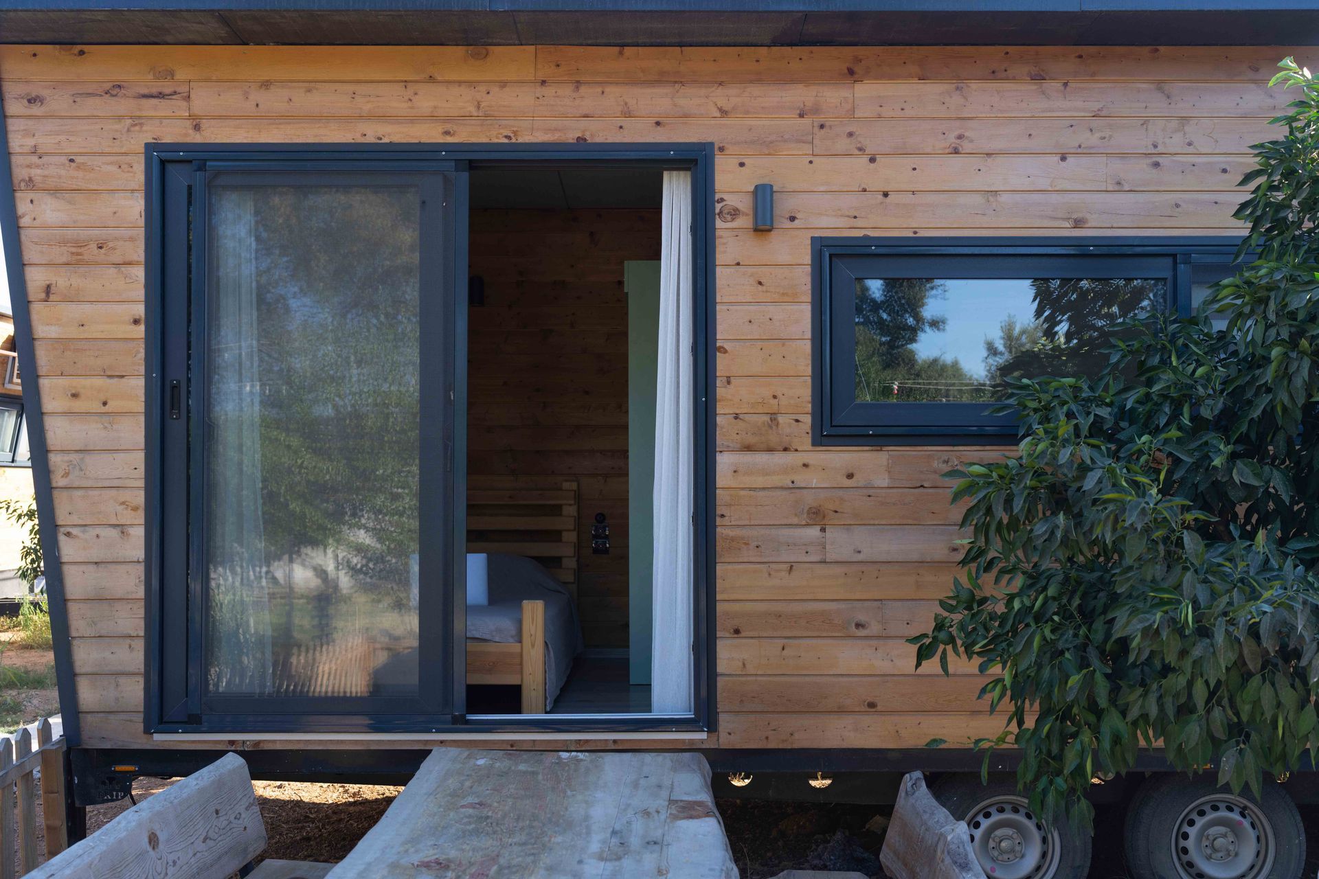 Modern wooden cabin entrance with open sliding glass door, window, and potted tree on the deck