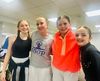 Four smiling dancers pose indoors, wearing a gray shirt, bright orange hoodie, and black outfits.