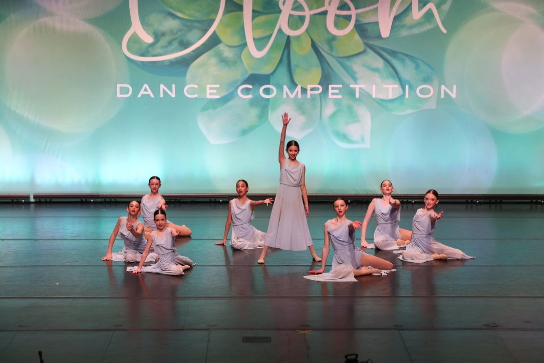 Dance troupe on stage under a “Bloom Dance Competition” backdrop, with one dancer standing center and others posed around her.