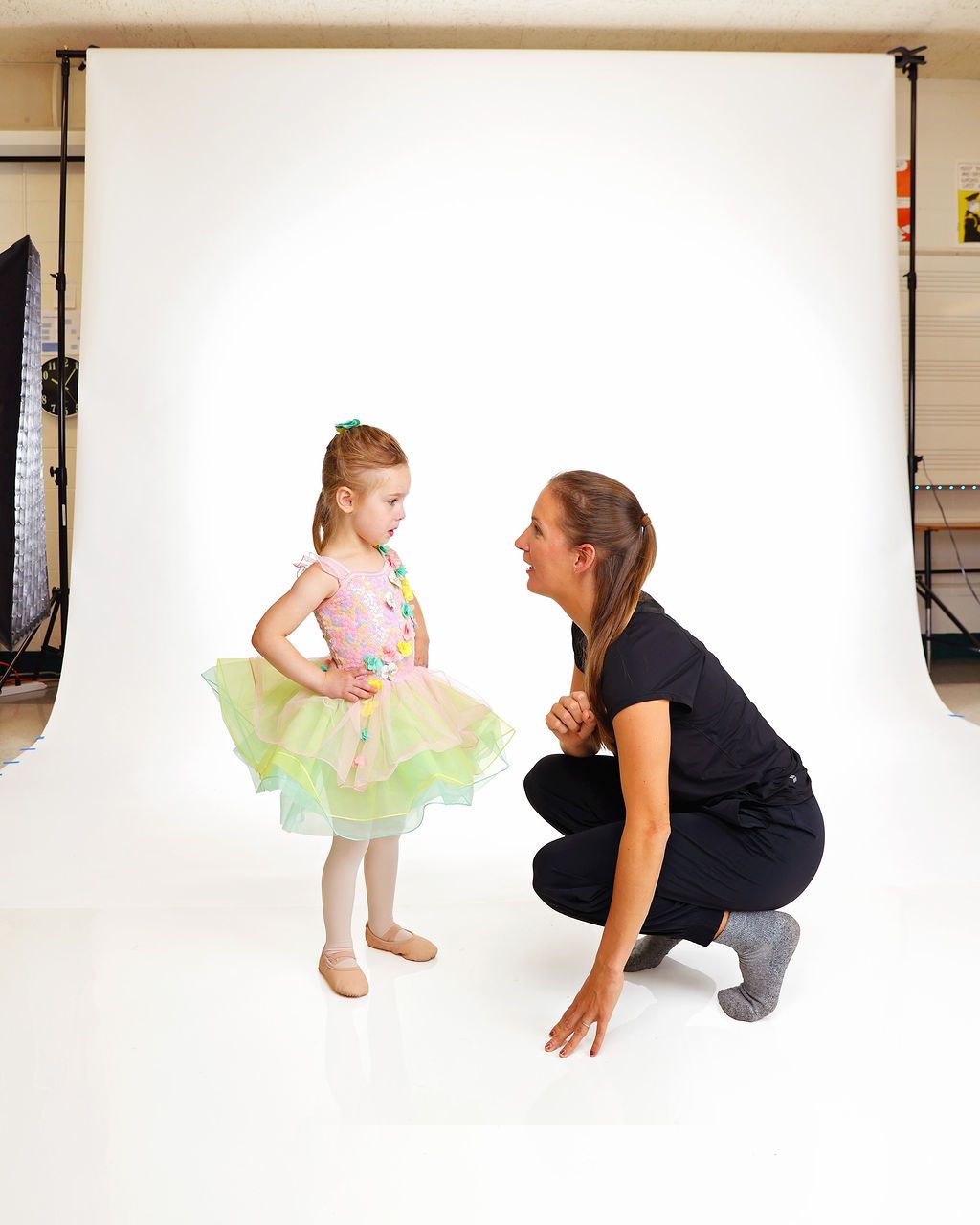 Woman crouching and smiling at a child in a colorful tutu against a white studio backdrop