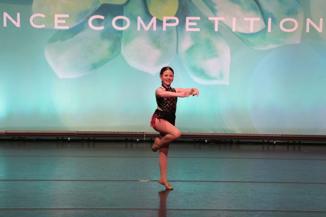 Young dancer in black costume posing onstage with one leg raised during a competition