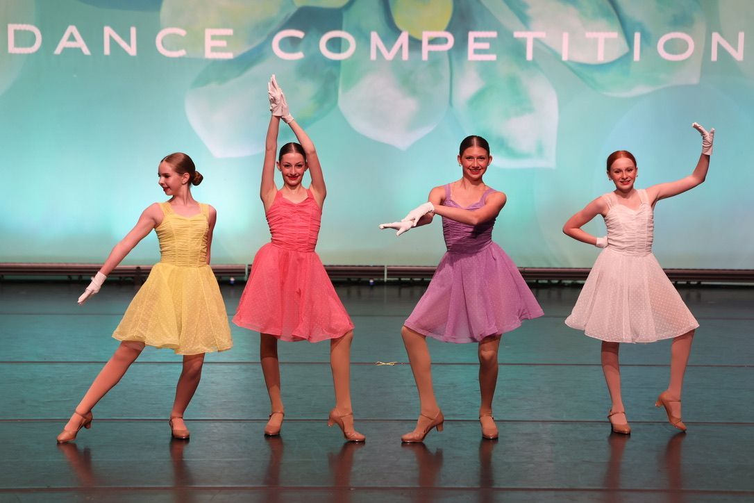 Four dancers in pastel dresses pose on a stage under “DANCE COMPETITION” signage.
