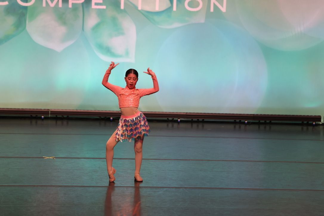 Young dancer in a pink top and patterned skirt poses on a stage with a competition backdrop