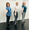 Three people pose in a dance studio, smiling and extending their arms toward the camera.