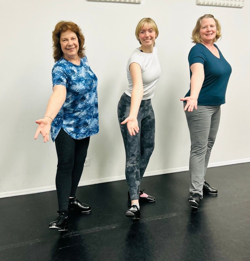 Three people posing in a dance studio, smiling with arms outstretched, wearing casual workout clothes.