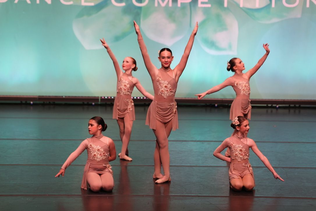 Five dancers in pink costumes pose onstage during a performance competition