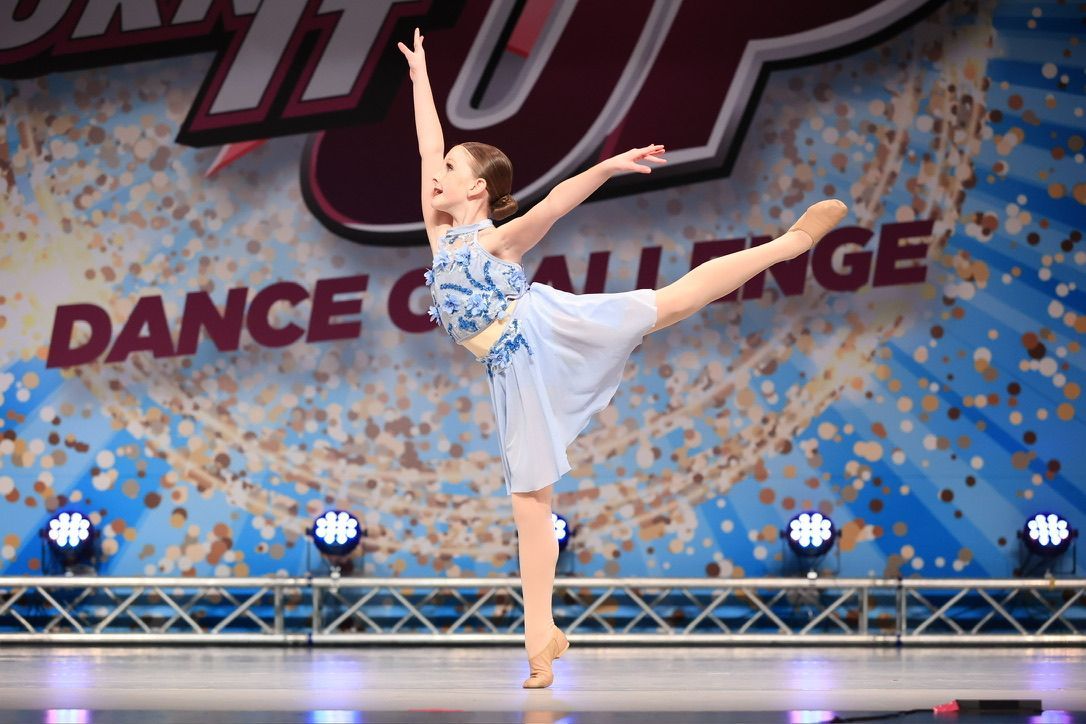 Young dancer in white costume performing a high leg extension on stage during a dance challenge