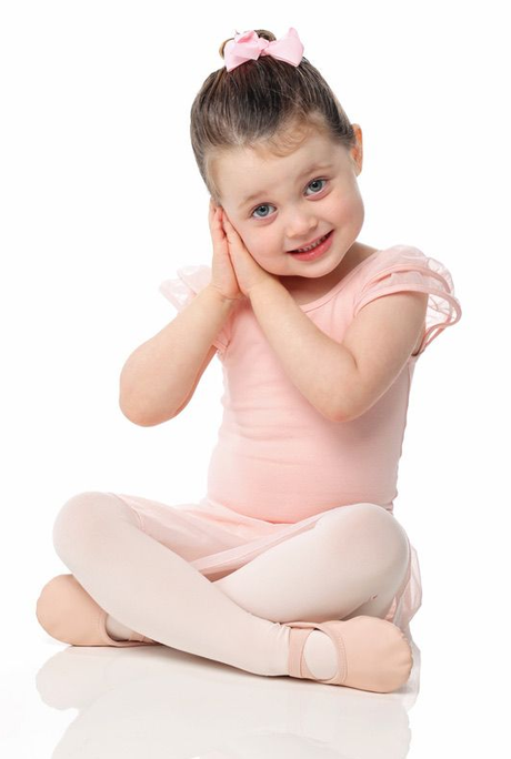 Young child in pink ballet outfit sitting cross-legged, smiling with hands pressed to cheek on a white background