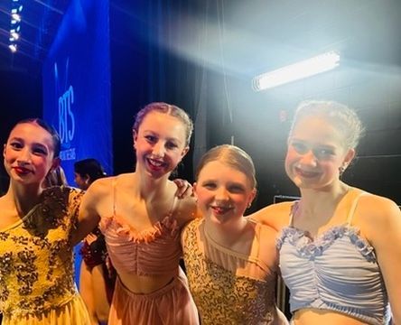 Four dancers in gold and blue costumes smiling onstage under bright lights