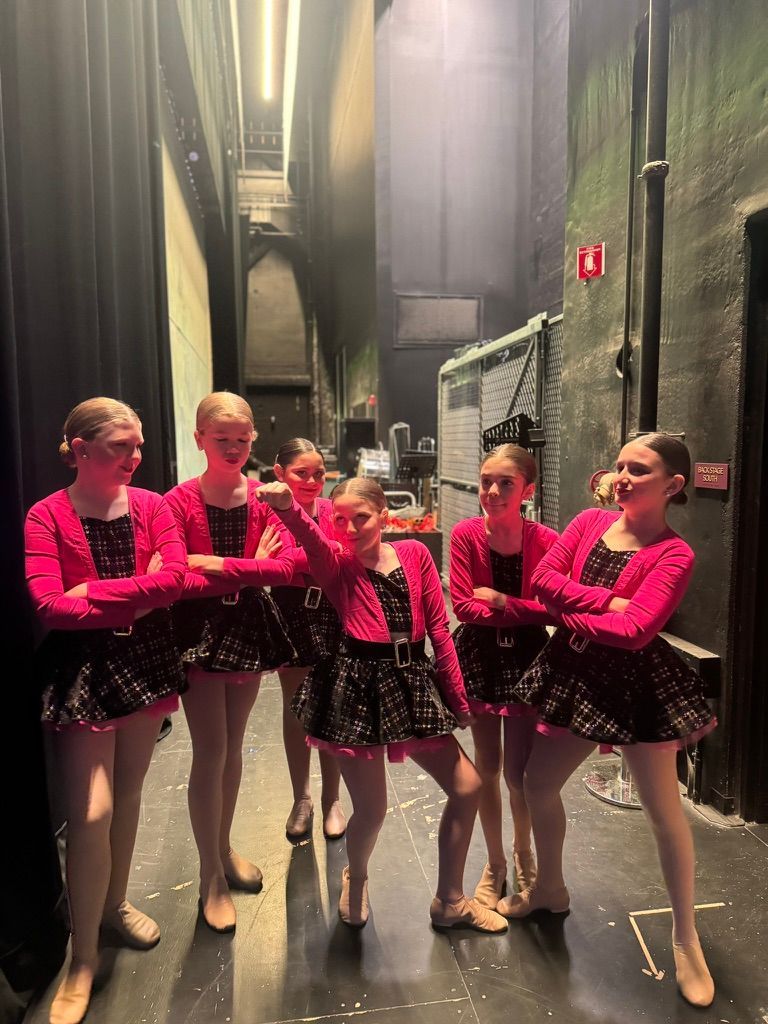 Five dancers in pink tops and black skirts pose backstage on a theater set