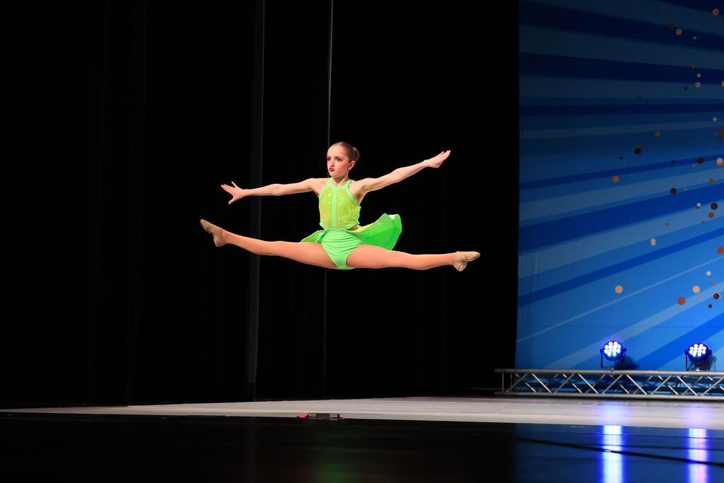 Gymnast in bright green leotard performing a split leap on a stage
