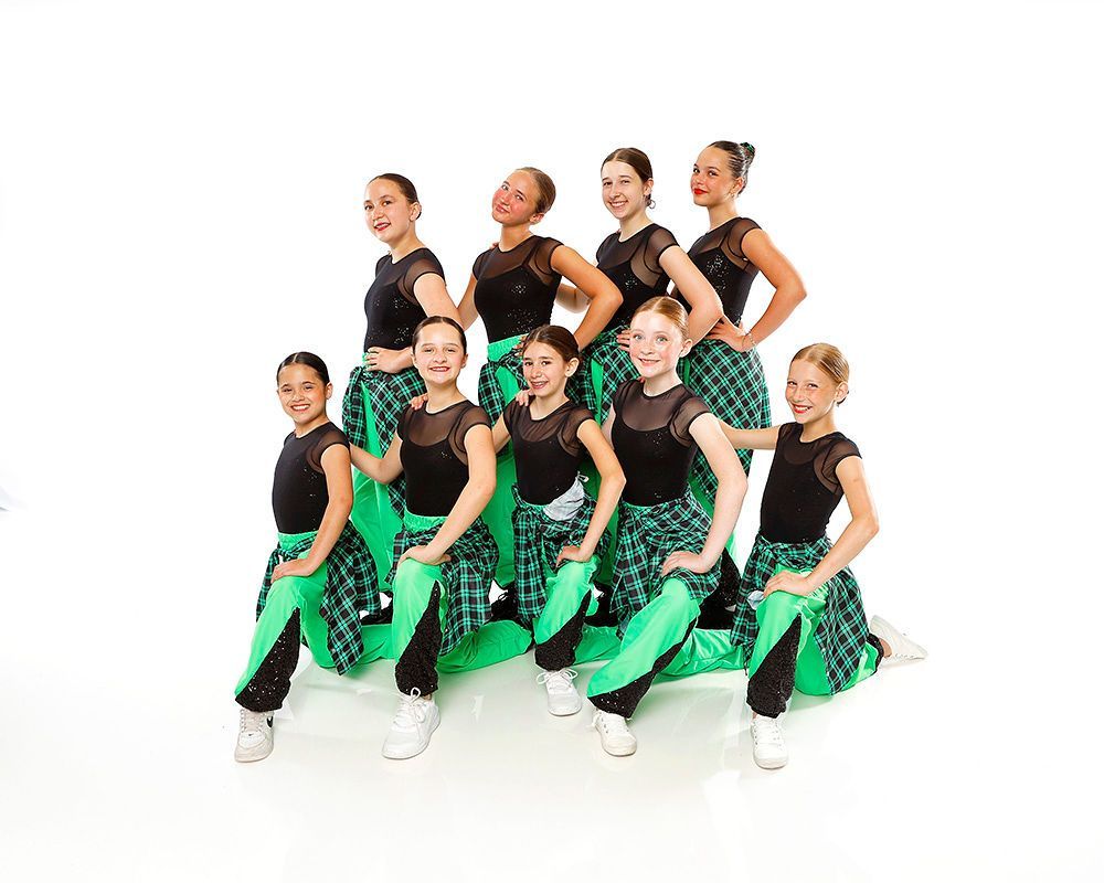 Group of dancers in black and green costumes posed in two rows against a white background