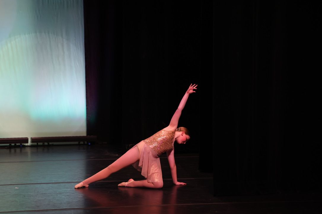 Dancer in a pink costume poses on a dark stage, one arm raised and one leg extended.