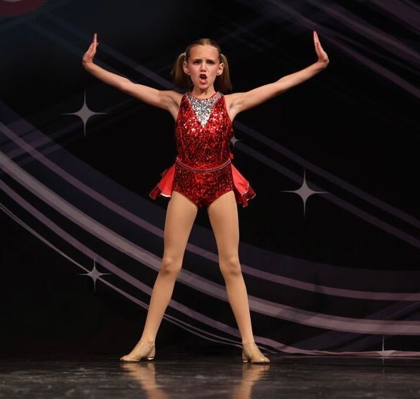 Young dancer in a red sequined costume posing with arms raised on stage