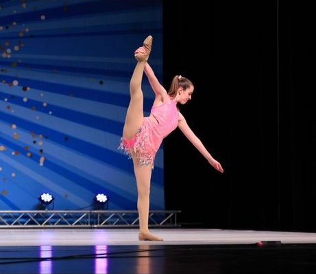 Young dancer in a pink costume balances in a high leg hold on a lit stage