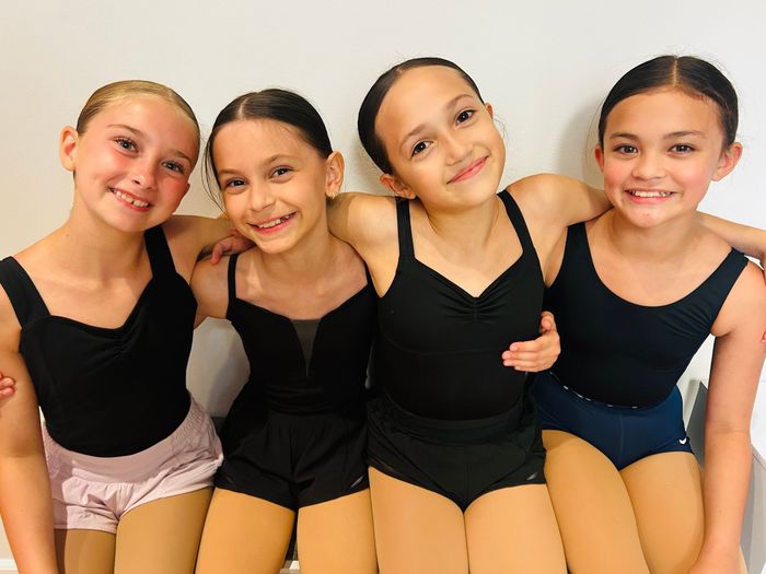 Four young dancers in black leotards and shorts smile with arms around each other against a light background
