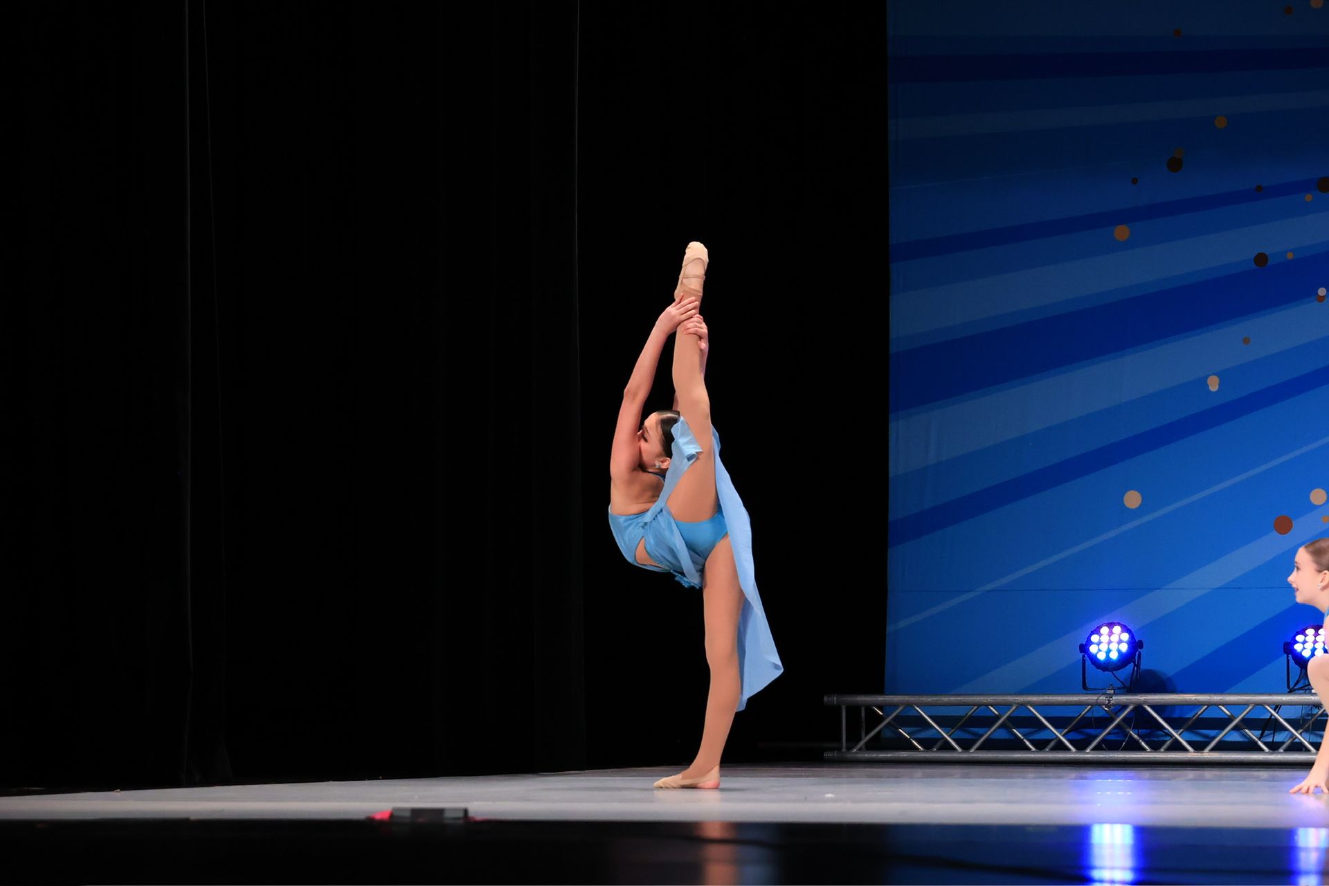 Dancer in blue leotard performs a high leg hold onstage under blue lighting.