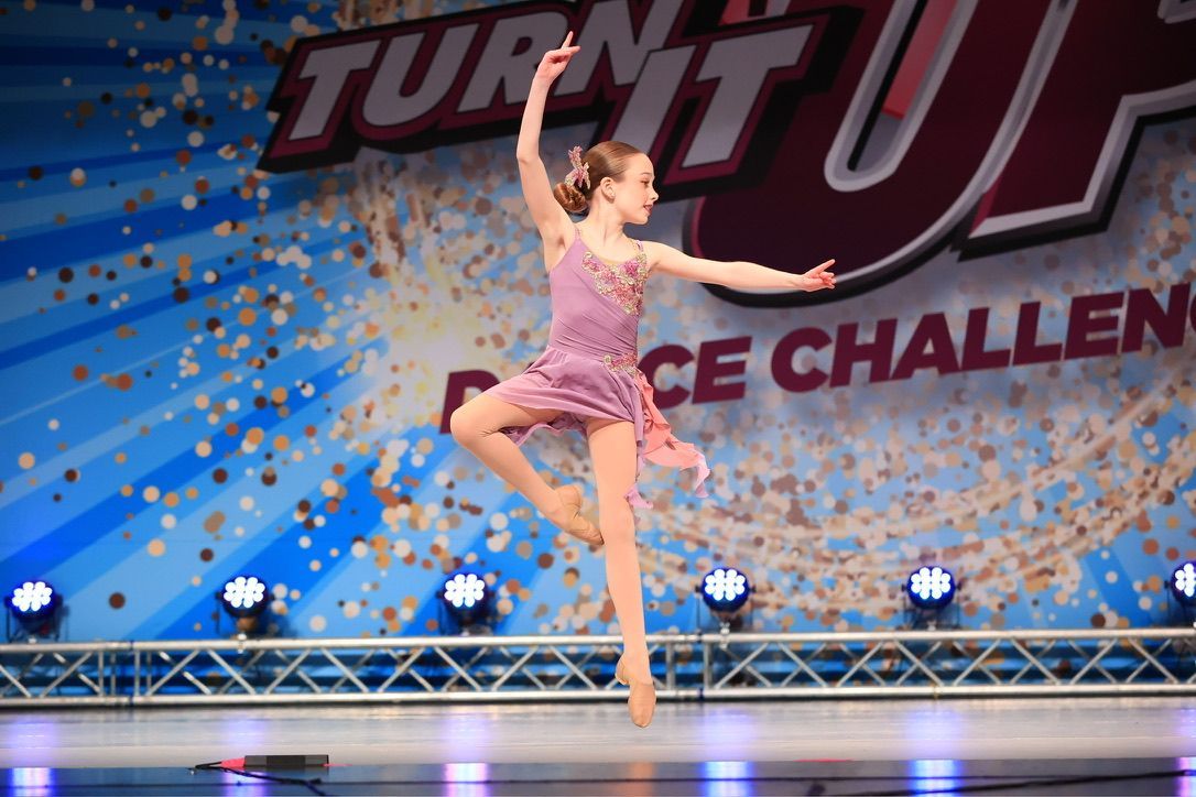Young figure skater in a pink costume performing on stage with TURN IT UP backdrop