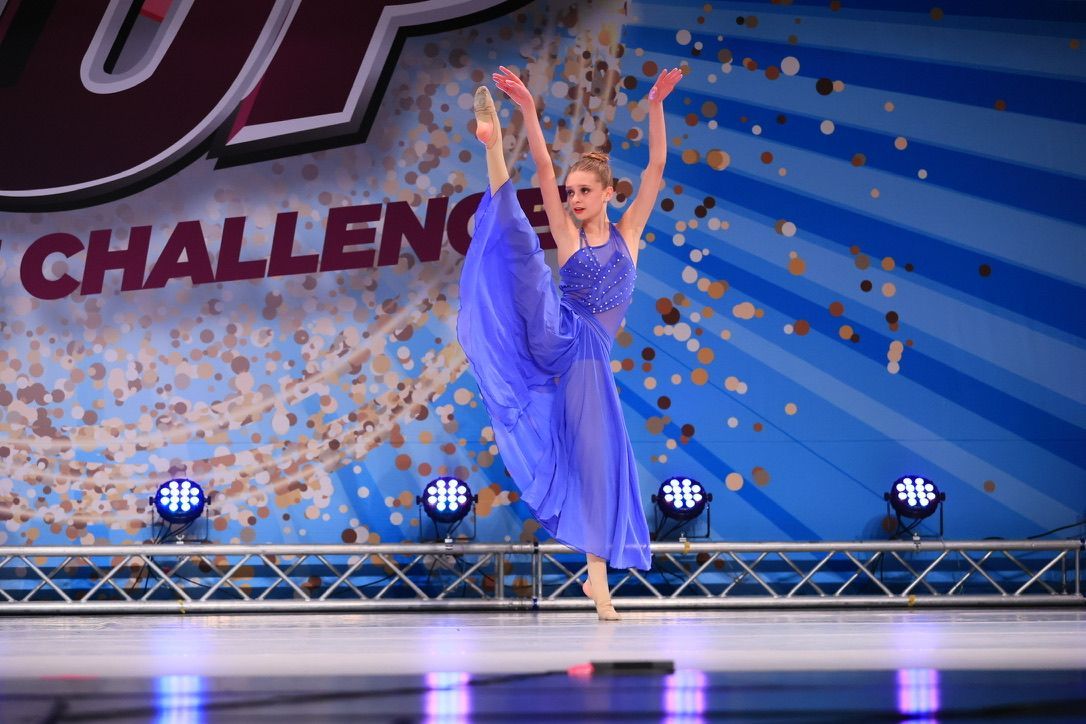 Dancer in a blue costume performing on stage with arms raised under blue lights