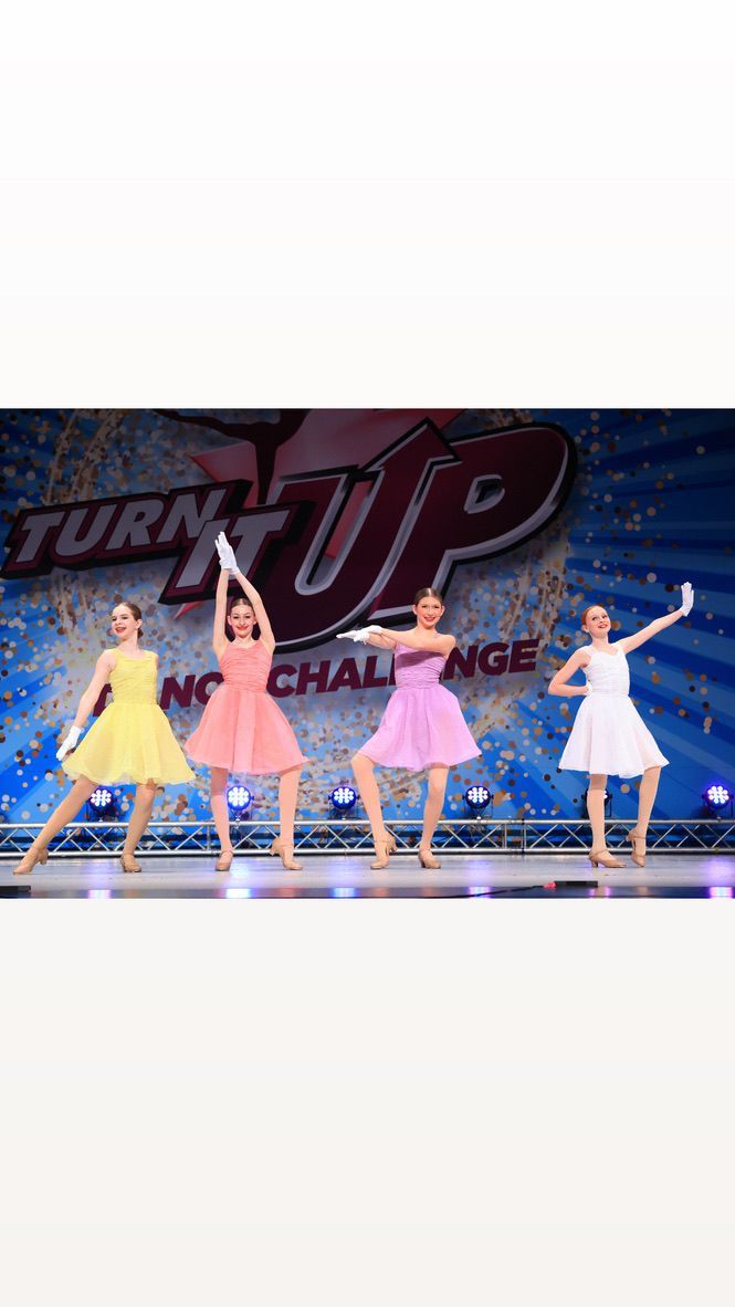 Four dancers perform onstage in yellow, orange, pink, and white dresses under a “Turn It Up Challenge” backdrop