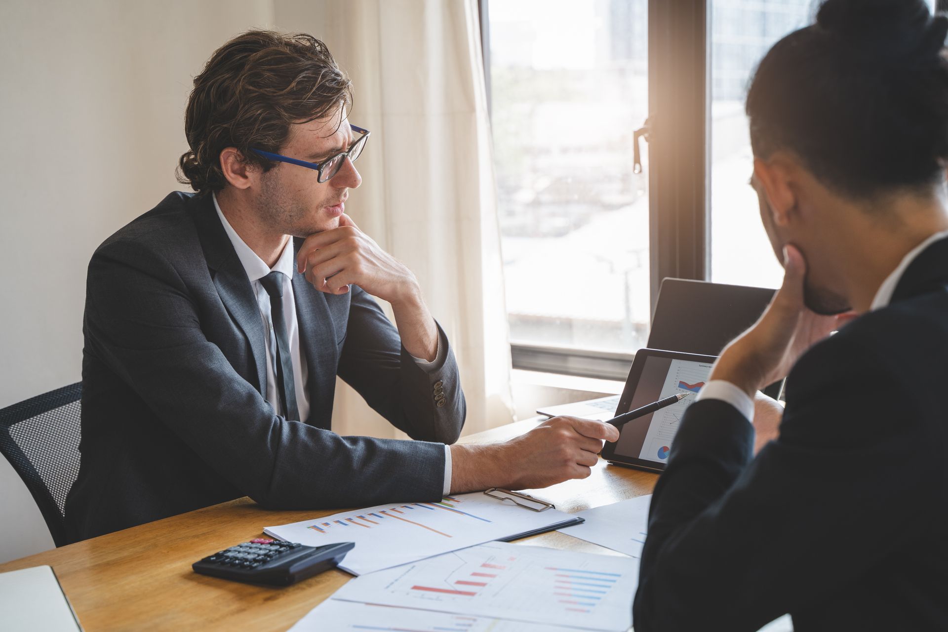Two people in suits looking at a tablet, pointing at charts, and discussing data at a desk.
