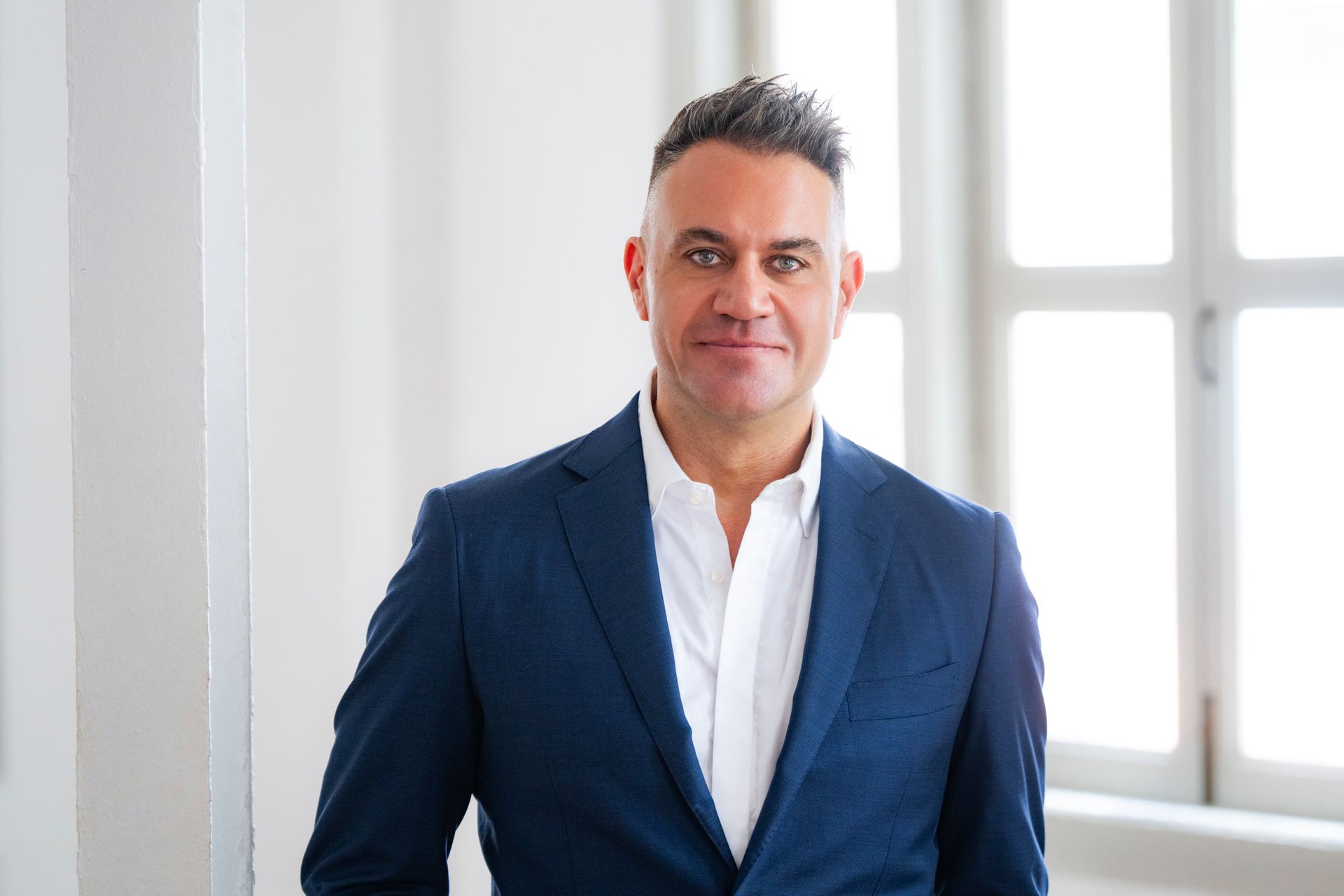 Man in suit speaks at a meeting, leaning on a table near two colleagues.